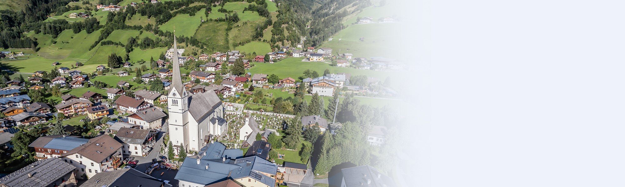 Luftaufnahme von Rauris mit der markanten Kirche im Zentrum, im Hintergrund grüne Hügel und verstreute Häuser